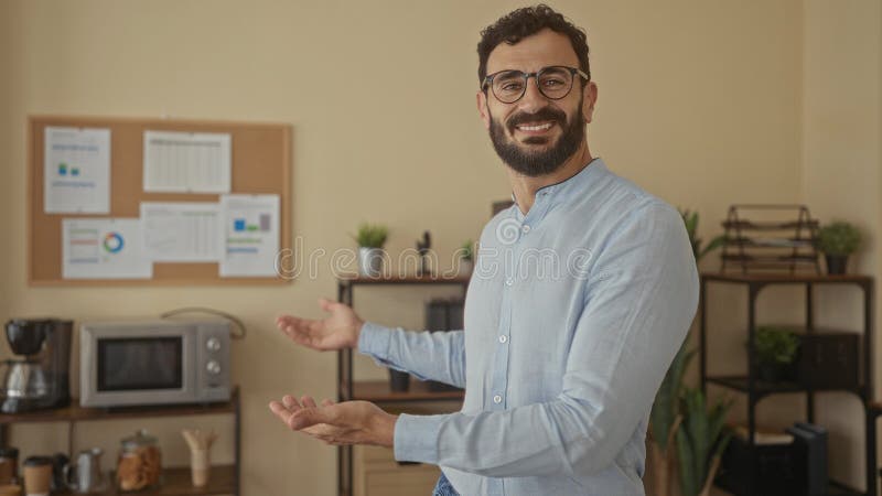 Hispanic man with beard smiling and gesturing in a modern office with charts on a corkboard, offering a warm and welcoming stock images