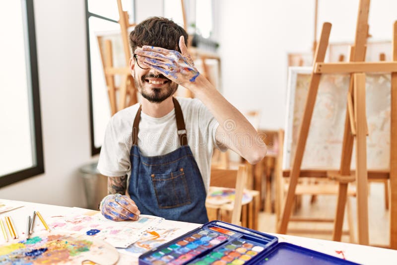 Hispanic man with beard at art studio smiling and laughing with hand on face covering eyes for surprise. blind concept. Hispanic man with beard at art studio smiling and laughing with hand on face covering eyes for surprise. blind concept