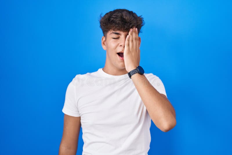 Hispanic teenager standing over blue background yawning tired covering half face, eye and mouth with hand