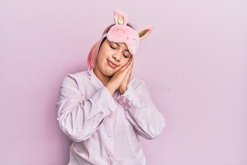 Hispanic woman with pink hair wearing sleep mask and pajama sleeping tired dreaming and posing with hands together while smiling