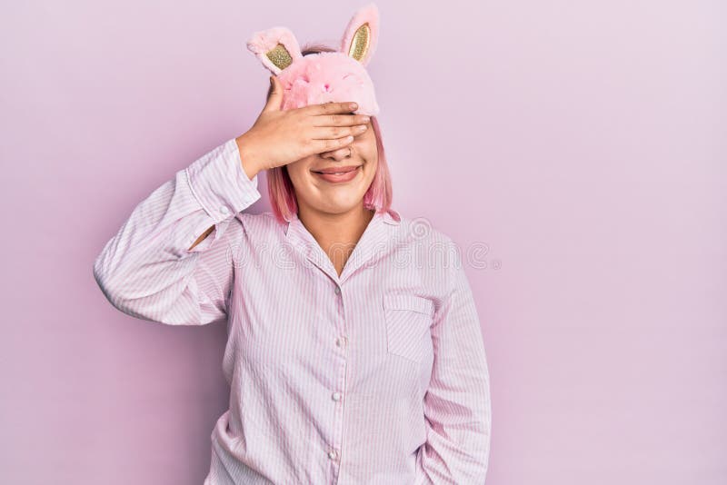 Hispanic woman with pink hair wearing sleep mask and pajama smiling and laughing with hand on face covering eyes for surprise