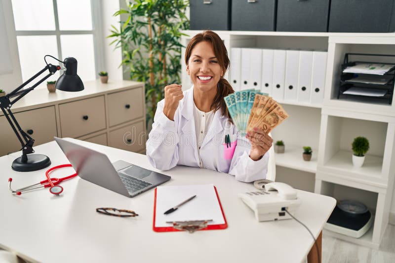 Hispanic doctor woman holding brazilian reals banknotes screaming proud, celebrating victory and success very excited with raised arms. Hispanic doctor woman holding brazilian reals banknotes screaming proud, celebrating victory and success very excited with raised arms