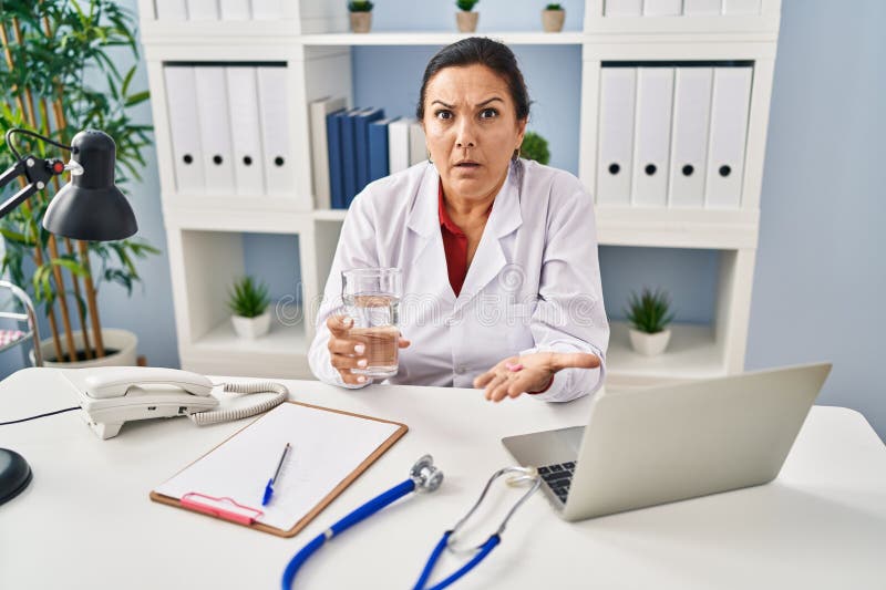 Hispanic mature doctor woman holding pill and glass of water in shock face, looking skeptical and sarcastic, surprised with open mouth. Hispanic mature doctor woman holding pill and glass of water in shock face, looking skeptical and sarcastic, surprised with open mouth