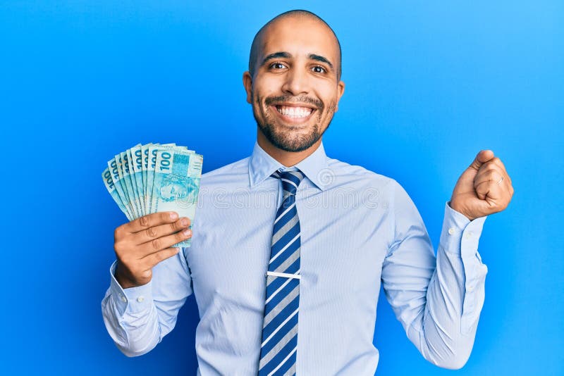 Hispanic adult man holding 100 brazilian real banknotes screaming proud, celebrating victory and success very excited with raised arm. Hispanic adult man holding 100 brazilian real banknotes screaming proud, celebrating victory and success very excited with raised arm