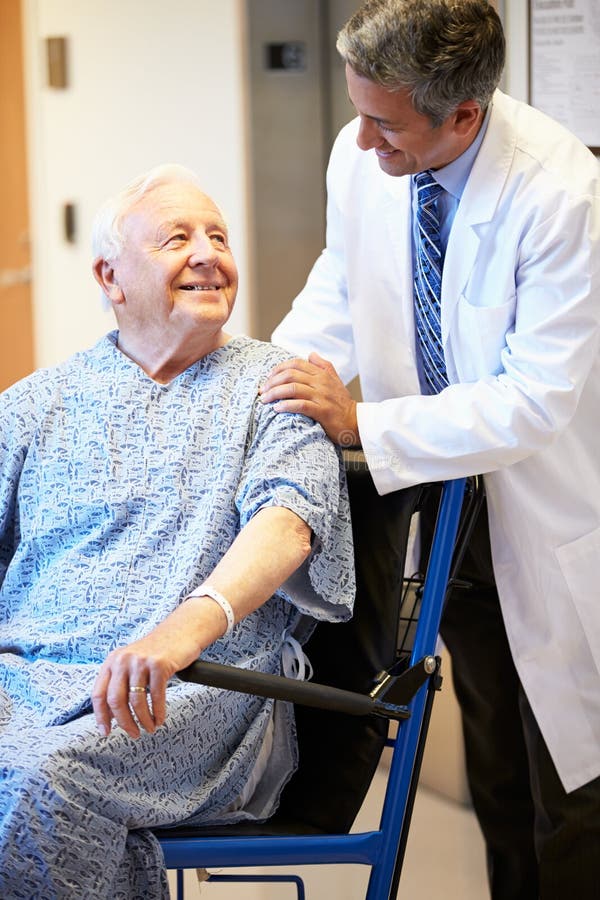 Senior Male Patient Being Pushed In Wheelchair By Doctor Looking At Each Other Smiling. Senior Male Patient Being Pushed In Wheelchair By Doctor Looking At Each Other Smiling