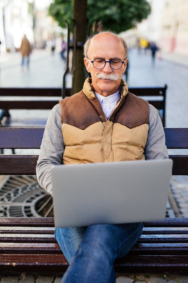 Hogere mens die aan zijn laptop zitting op de bank in de stadsstraat werken royalty-vrije stock foto's Hogere mens die aan zijn laptop zitting op de bank in de stadsstraat werken royalty-vrije stock foto's