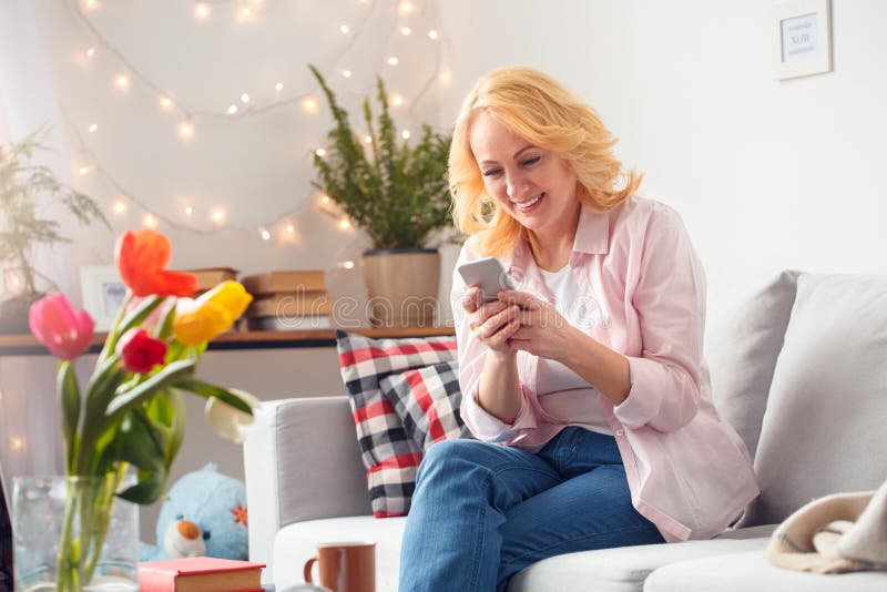 Hogere vrouw die thuis het doorbladeren blije smartphone zitten stock foto Hogere vrouw die thuis het doorbladeren blije smartphone zitten stock foto