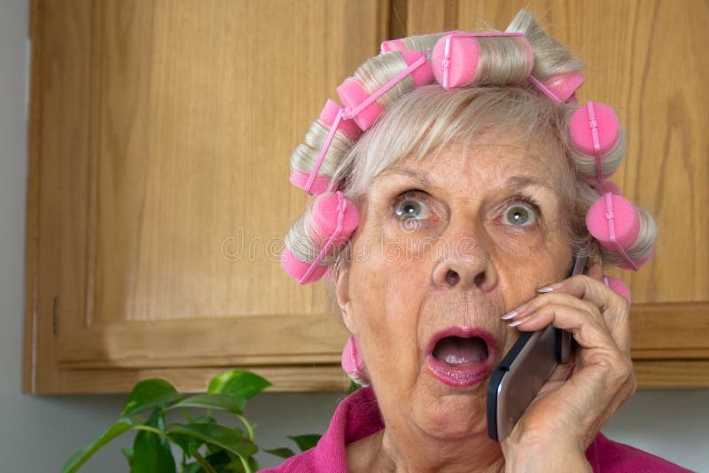 Shocked senior woman in pink curlers in her kitchen listens on her cell phone. Shocked senior woman in pink curlers in her kitchen listens on her cell phone.