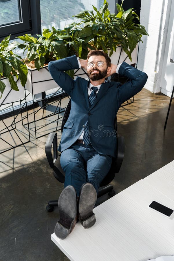 hohe Winkelsicht des lächelnden bärtigen Geschäftsmannes, der mit den Beinen auf Tabelle und Händen hinter Kopf sich entspannt lizenzfreies stockbild hohe Winkelsicht des lächelnden bärtigen Geschäftsmannes, der mit den Beinen auf Tabelle und Händen hinter Kopf sich entspannt lizenzfreies stockbild