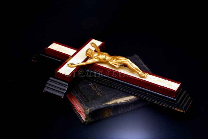 An old wooden cross with golden figure of christ is placed on top of an antique bible. With blue accent light on black background. An old wooden cross with golden figure of christ is placed on top of an antique bible. With blue accent light on black background.