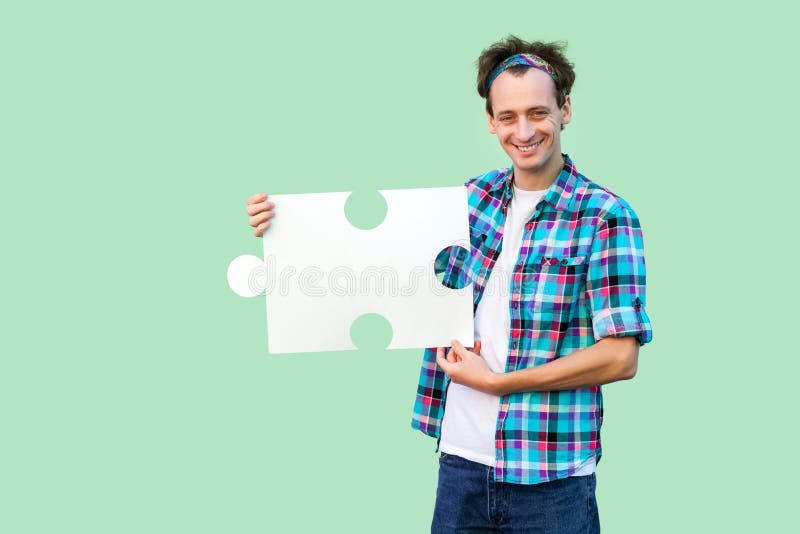 Hombre adulto joven hermoso feliz en la camisa a cuadros que coloca y que lleva a cabo el pedazo grande de rompecabezas, mirando imagen de archivo libre de regalías Hombre adulto joven hermoso feliz en la camisa a cuadros que coloca y que lleva a cabo el pedazo grande de rompecabezas, mirando imagen de archivo libre de regalías
