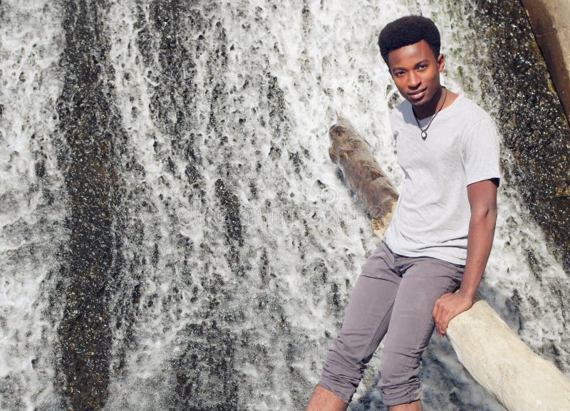 Hombre africano joven que se sienta en tronco de árbol en cascada del río imágenes de archivo libres de regalías Hombre africano joven que se sienta en tronco de árbol en cascada del río imágenes de archivo libres de regalías