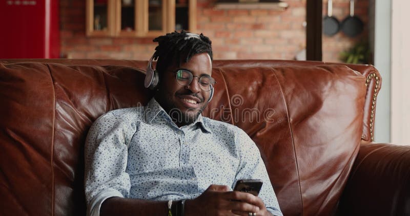 Hombre africano sonriente en audífonos se relaja en un sofá usando teléfono metrajes