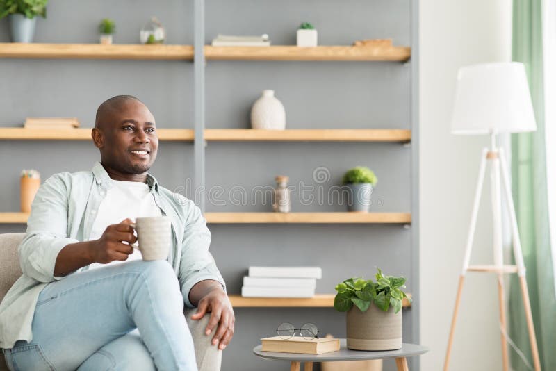 Hombre afroamericano sonriente y reflexivo tomando café sentado en un sillón en una sala de estar espacio libre interior imagen de archivo