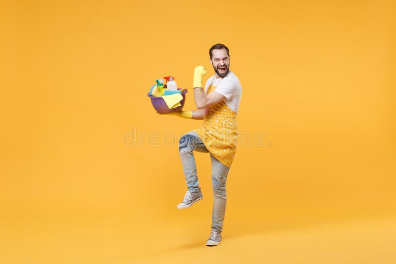 Hombre alegre ama de casa en guantes de delantal sostener botellas detergentes lavadoras de cuencas haciendo las tareas domésticas foto de archivo