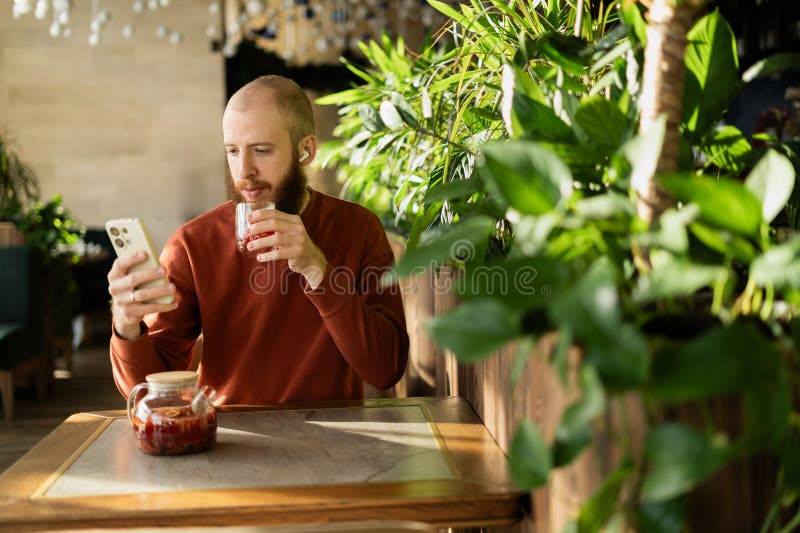 Hombre apuesto con barba tomando té leyendo noticias y medios sociales usando teléfono móvil en café verde. fotos de archivo libres de regalías