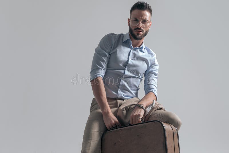 Hombre apuesto y sin afeitar con gafas sosteniendo maleta y posando foto de archivo