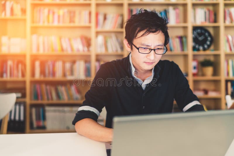 Hombre asiático joven que trabaja en oficina o biblioteca del ordenador portátil en casa con la cara seria, estante con el fondo fotografía de archivo Hombre asiático joven que trabaja en oficina o biblioteca del ordenador portátil en casa con la cara seria, estante con el fondo fotografía de archivo