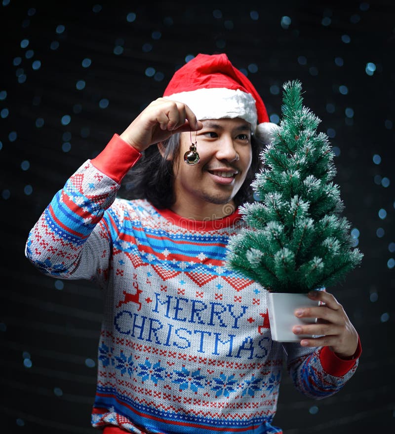Hombre asiático que lleva Santa Hat y el suéter de la Navidad que se considera pequeños imagen de archivo libre de regalías Hombre asiático que lleva Santa Hat y el suéter de la Navidad que se considera pequeños imagen de archivo libre de regalías