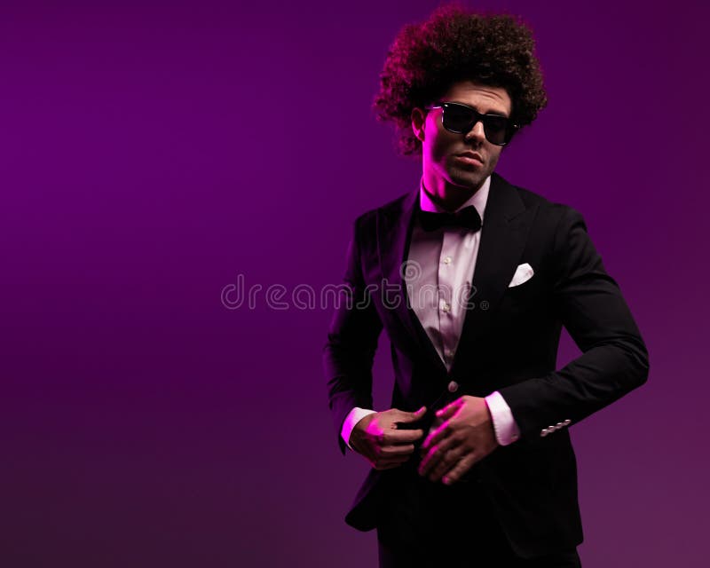 Hombre atractivo con gafas de sol y pelo rizado ajustando esmoquin negro fotografía de archivo