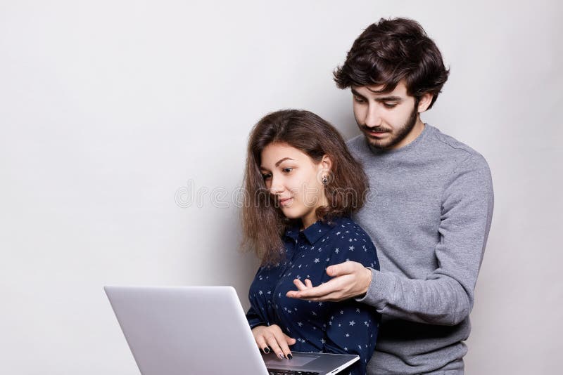 Hombre barbudo elegante y mujer bonita que se colocan de lado delante del ordenador portátil abierto y que discuten algo, mirando foto de archivo Hombre barbudo elegante y mujer bonita que se colocan de lado delante del ordenador portátil abierto y que discuten algo, mirando foto de archivo