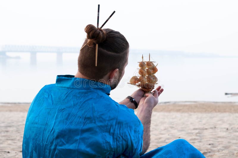 Hombre barbudo en kimono azul con el modelo de nave de la tenencia del bollo del pelo con las conchas marinas delante del río imagenes de archivo Hombre barbudo en kimono azul con el modelo de nave de la tenencia del bollo del pelo con las conchas marinas delante del río imagenes de archivo
