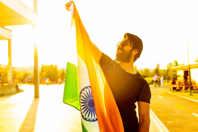 Hombre barbudo indio guapo en camiseta de algodón negro enarbolando la bandera india en el parque de verano en la luz dorada al at fotografía de archivo libre de regalías