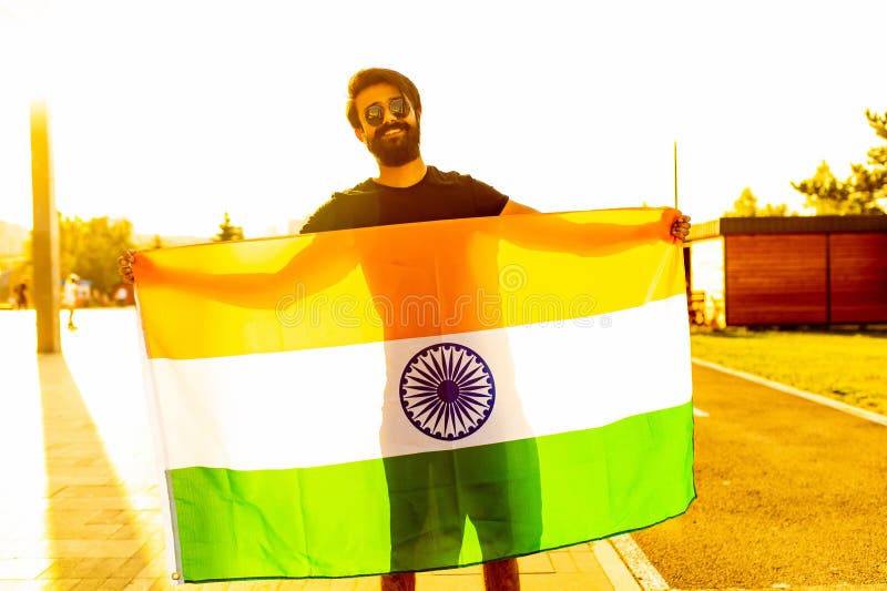 Hombre barbudo indio guapo en camiseta de algodón negro enarbolando la bandera india en el parque de verano en la luz dorada al at imagen de archivo libre de regalías