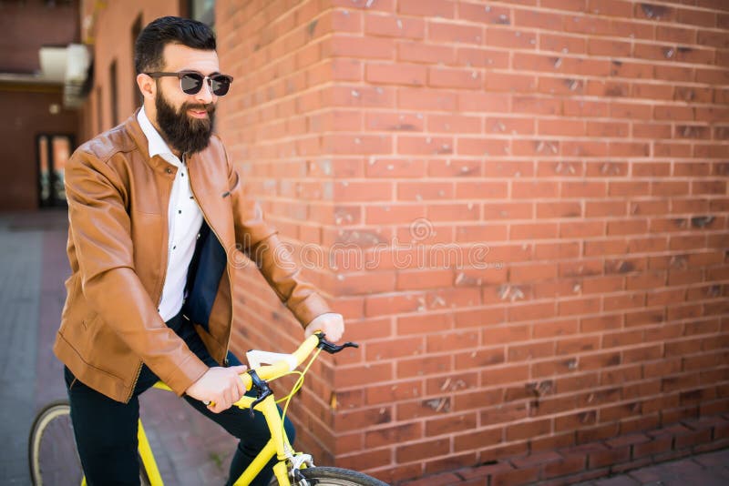 Hombre barbudo joven hermoso mientras que se opone cerca de su bicicleta a la pared de ladrillo fotografía de archivo Hombre barbudo joven hermoso mientras que se opone cerca de su bicicleta a la pared de ladrillo fotografía de archivo