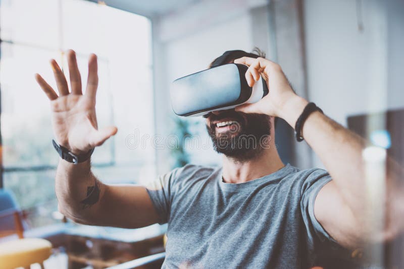 Hombre barbudo joven que lleva los vidrios de la realidad virtual en estudio coworking moderno del diseño interior Smartphone usa fotografía de archivo Hombre barbudo joven que lleva los vidrios de la realidad virtual en estudio coworking moderno del diseño interior Smartphone usa fotografía de archivo