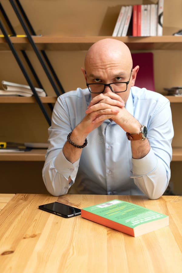 Hombre calvo serio con anteojos esta sentado a la mesa imagen de archivo