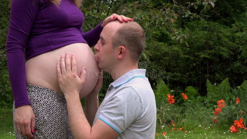 Hombre cariñoso del marido que frota ligeramente y que se besa su panza embarazada de la mujer de la esposa en jardín