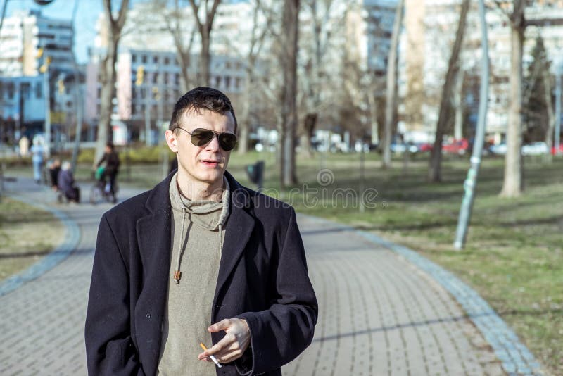 Hombre casual joven del fumador con las gafas de sol en el cigarrillo que fuma de la capa negra afuera en el parque imagen de archivo libre de regalías Hombre casual joven del fumador con las gafas de sol en el cigarrillo que fuma de la capa negra afuera en el parque imagen de archivo libre de regalías