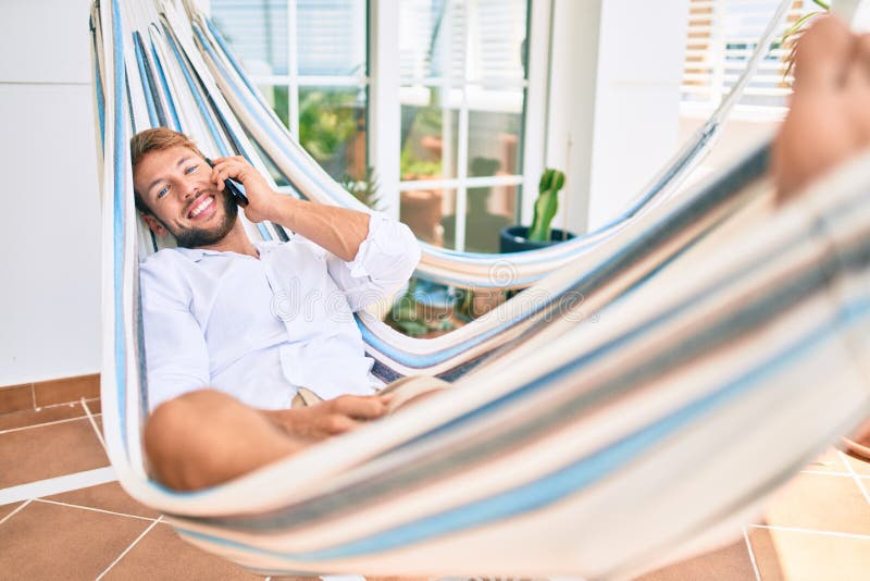 Hombre caucásico apuesto sonriendo feliz descansando en una hamaca en la terraza hablando por teléfono foto de archivo libre de regalías