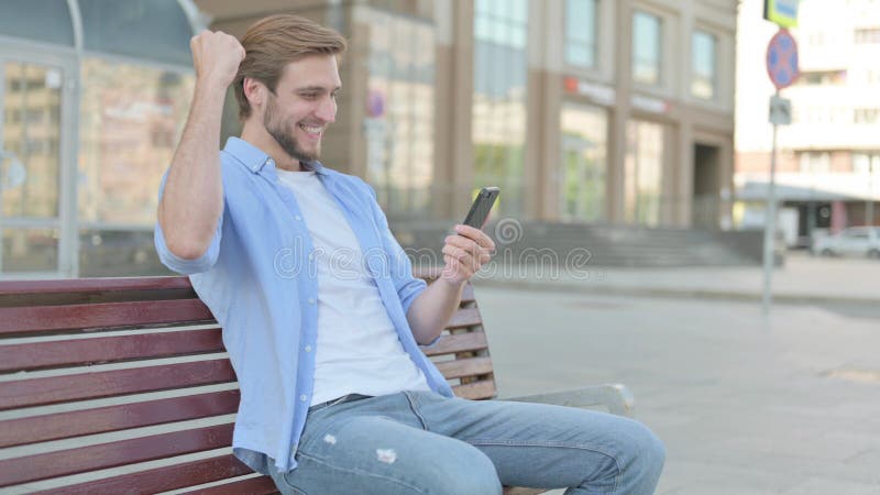Hombre celebrando el éxito en línea en el smartphone mientras estaba sentado al aire libre en el banco imágenes de archivo libres de regalías