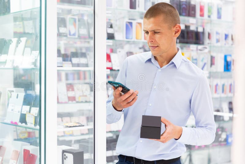 Smiling elegant young man standing in tech store with new smart phone in his hands. Smiling elegant young man standing in tech store with new smart phone in his hands