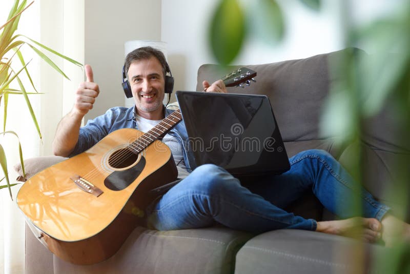 Hombre con auriculares de guitarra y gesto de laptop ok en sofá fotos de archivo libres de regalías