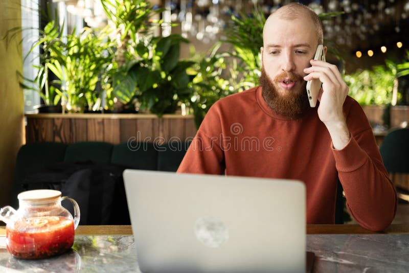 Hombre con barba en suéter hablando en un empresario de telefonía celular que trabaja en un café usando una computadora portátil c imágenes de archivo libres de regalías