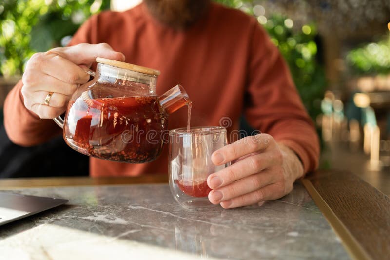Hombre con barba usando suéter vertiendo té de una tetera de vidrio a una taza en la mesa en un café verde. fotografía de archivo libre de regalías