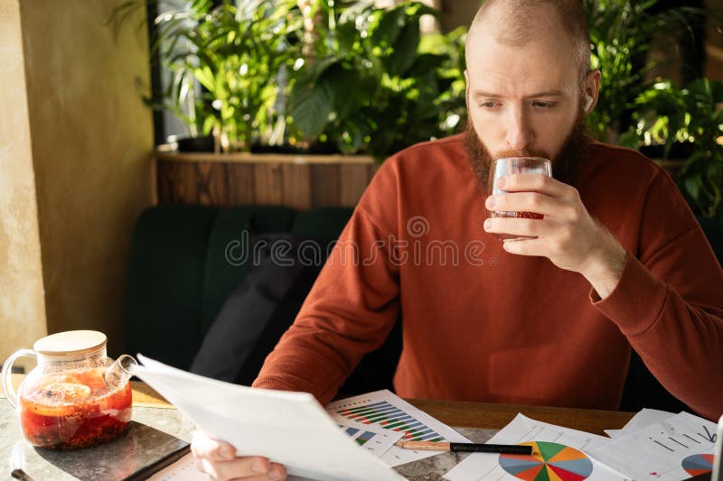 Hombre con barba y suéter cálido sostiene una factura y otros documentos tomando té centrado en el manejo de sus finanzas sentados foto de archivo libre de regalías