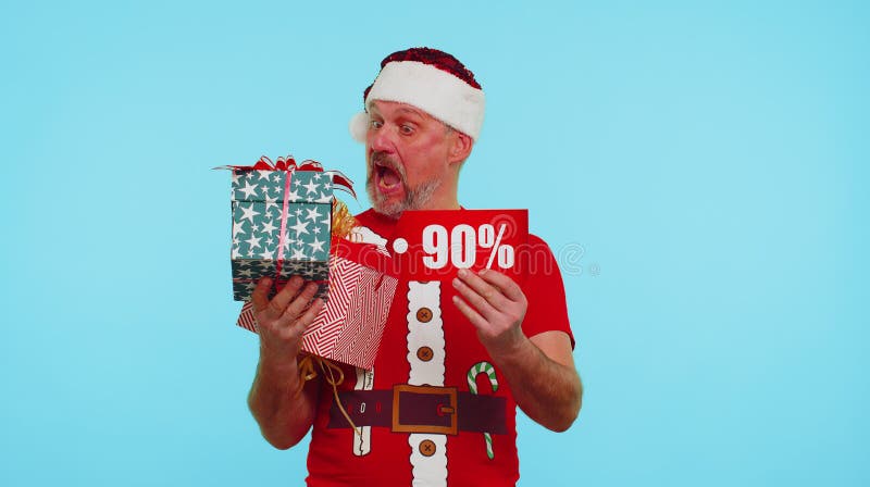 Senior man wears red New Year t-shirt showing gift box and 90 Percent discount inscriptions banner text. Black Friday. Advertising low prices for shopping. Blue studio wall background. Merry Christmas. Senior man wears red New Year t-shirt showing gift box and 90 Percent discount inscriptions banner text. Black Friday. Advertising low prices for shopping. Blue studio wall background. Merry Christmas