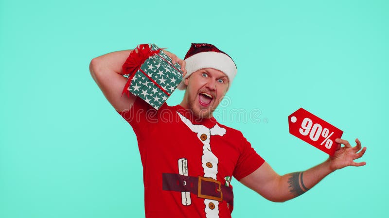 Young man wears red New Year t-shirt showing gift box and 90 Percent discount inscriptions banner text. Black Friday. Advertising low prices for shopping. Blue studio wall background. Merry Christmas. Young man wears red New Year t-shirt showing gift box and 90 Percent discount inscriptions banner text. Black Friday. Advertising low prices for shopping. Blue studio wall background. Merry Christmas