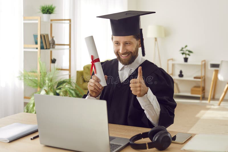 Hombre con traje de graduado sentado en un escritorio con diploma que muestra el pulgar hacia arriba durante una videollamada. foto de archivo