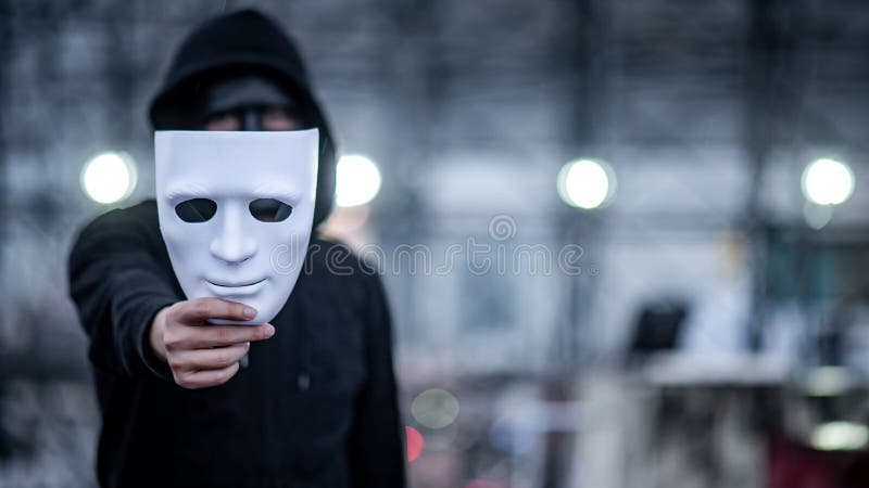 Mystery hoodie man with black mask holding white mask in his hand. Anonymous social masking or bipolar disorder concept. Focus on white mask. Mystery hoodie man with black mask holding white mask in his hand. Anonymous social masking or bipolar disorder concept. Focus on white mask