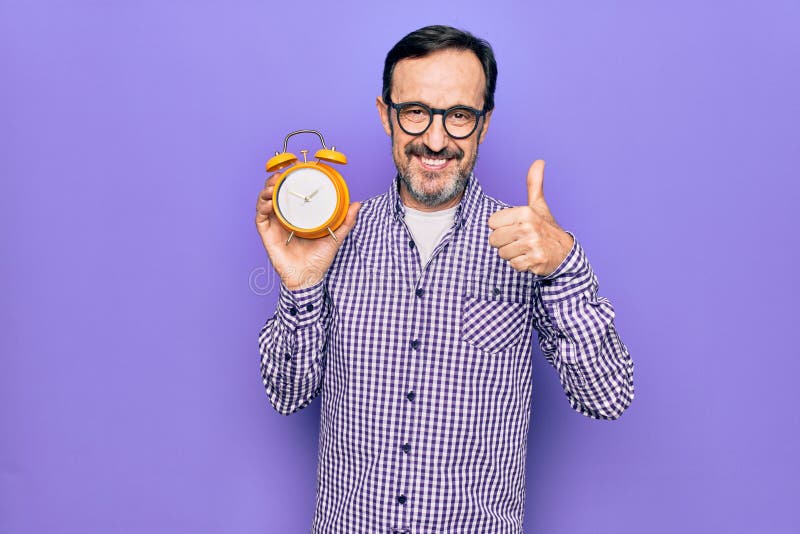Hombre de mediana edad con gafas sosteniendo vintage despertador sobre fondo púrpura aislado sonriendo feliz y pulgar positivo hac imagenes de archivo