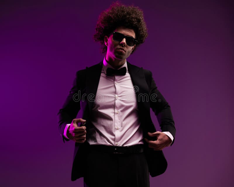 Hombre de moda fresco con pelo rizado y gafas de sol ajustando esmoquin negro fotografía de archivo