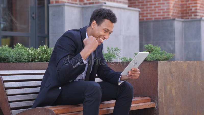 Hombre de negocios africano emocionado Celebrating Success en la tableta que se sienta en banco foto de archivo libre de regalías Hombre de negocios africano emocionado Celebrating Success en la tableta que se sienta en banco foto de archivo libre de regalías