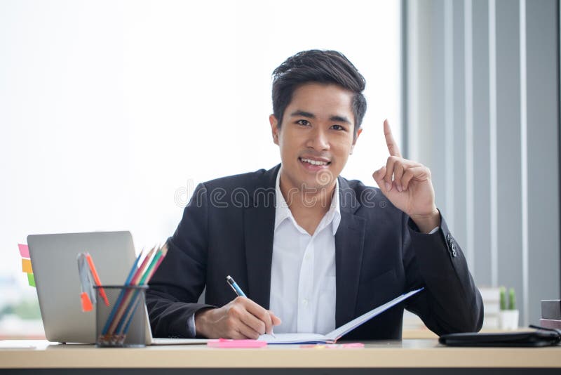 Hombre de negocios asiático joven sonriente que trabaja con el cuaderno en el escritorio y el finger que señalan para arriba en u foto de archivo libre de regalías Hombre de negocios asiático joven sonriente que trabaja con el cuaderno en el escritorio y el finger que señalan para arriba en u foto de archivo libre de regalías