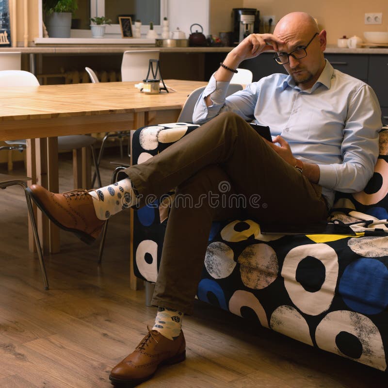 Hombre de negocios calvo en anteojos usando el teléfono móvil sentado en el sofá en la habitación. configuración de oficina domést foto de archivo libre de regalías