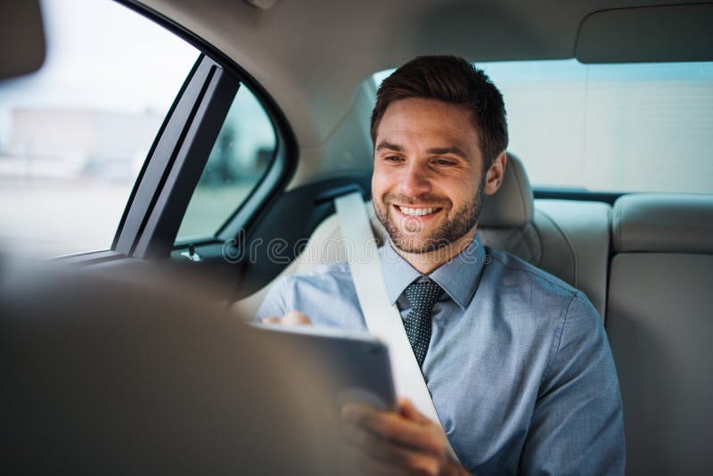 Hombre de negocios con la tableta que se sienta en asientos traseros en el coche, trabajando foto de archivo libre de regalías Hombre de negocios con la tableta que se sienta en asientos traseros en el coche, trabajando foto de archivo libre de regalías